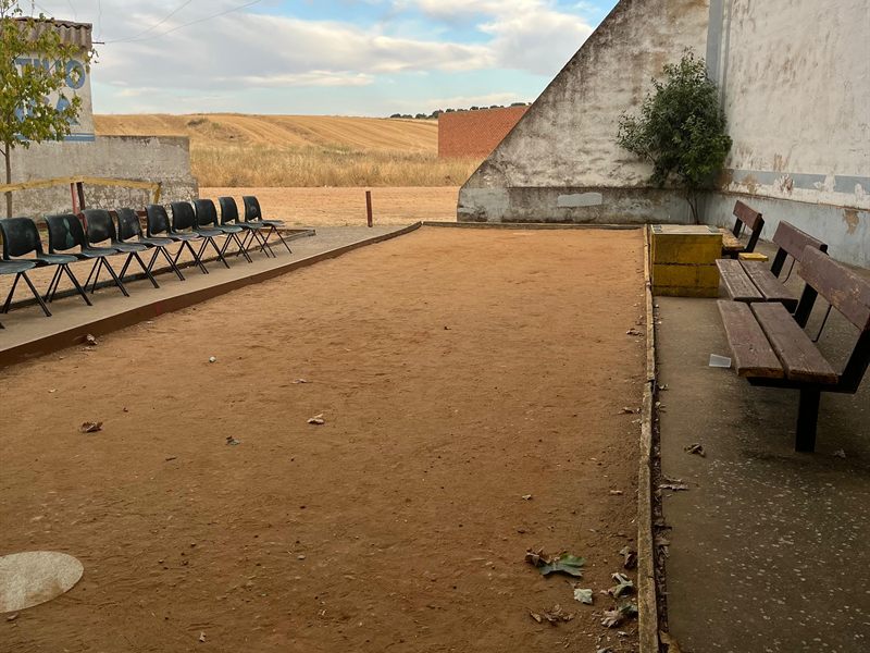 Pista de petanca de tierra, con asientos corridos a un lado y los campos de cereal al fondo.