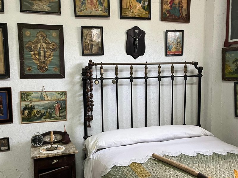 Alcoba recreada en el museo, con cama de hierro forjado y estampas religiosas en la pared